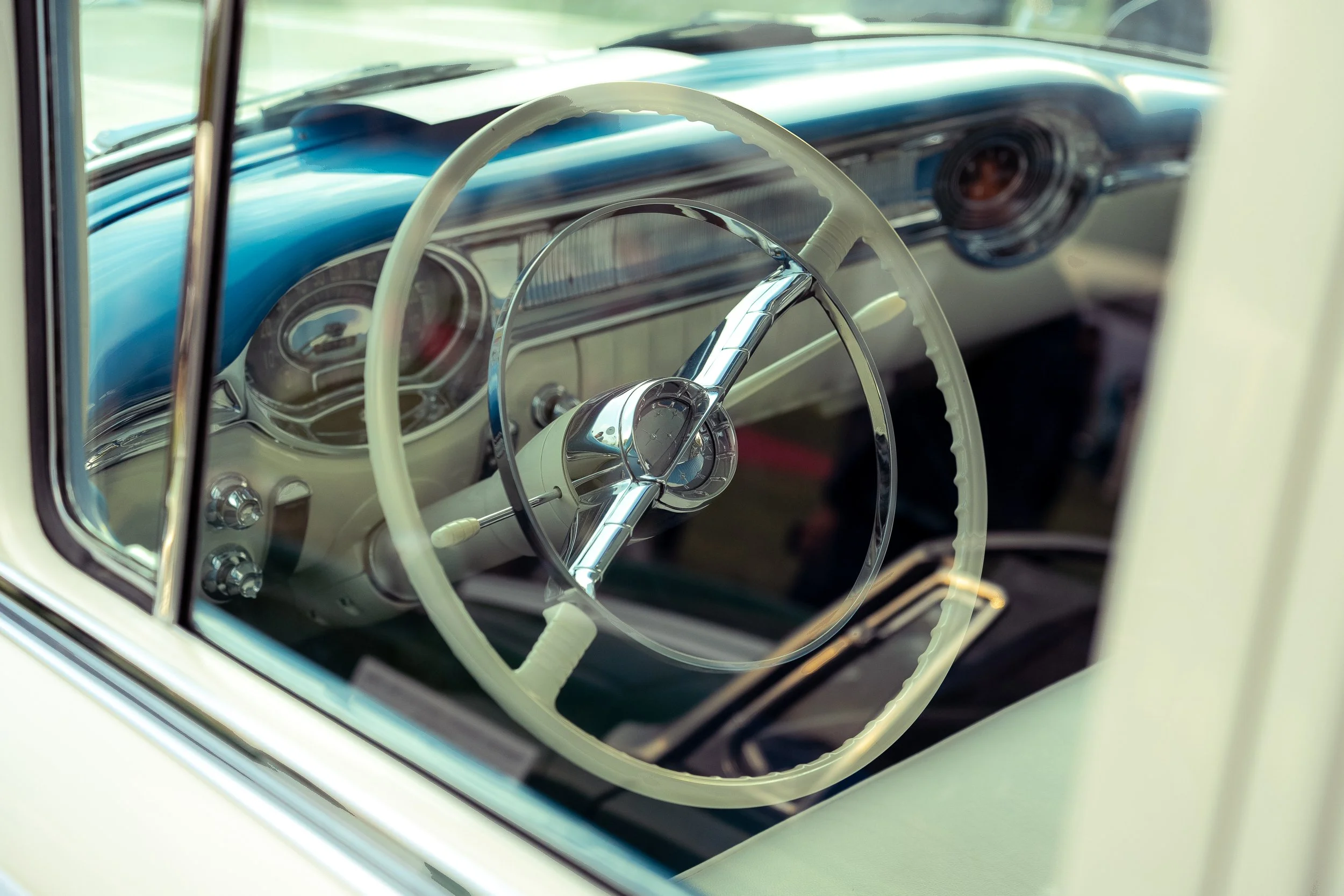 Ivory steering wheel and chrome horn ring seen through the open window of a blue and cream classic car