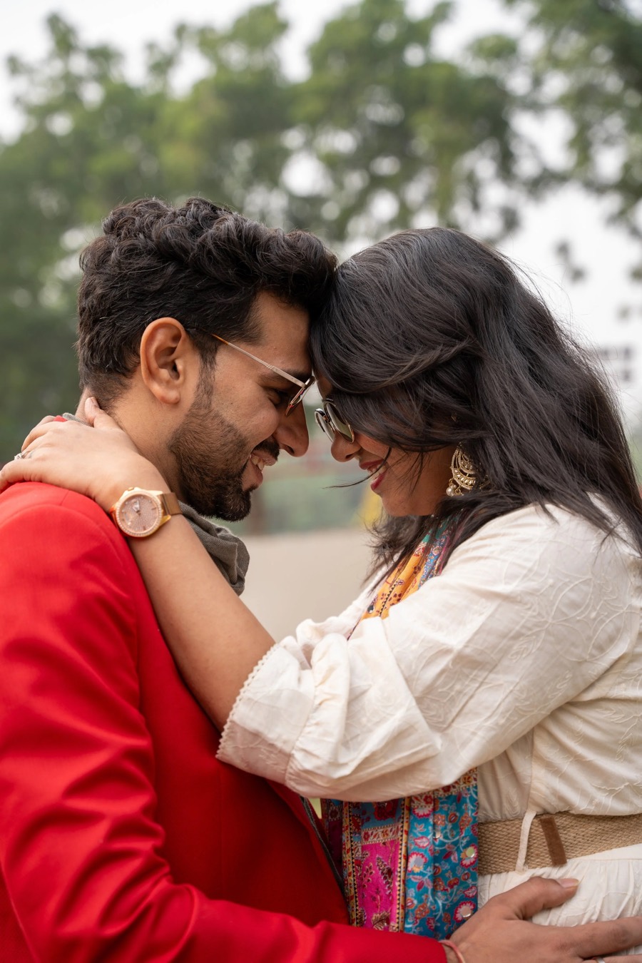 Foreheads pressed together outdoors, a red jacket and a white one, arms wrapped close against green trees