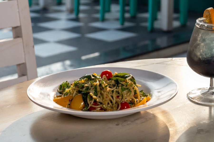 Herbed pasta with cherry tomatoes on a marble cafe table, iced drink alongside and checked floor behind