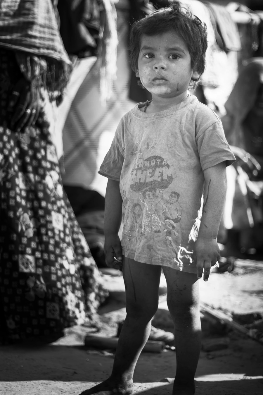 Child in a graphic T-shirt standing still in a busy market lane, figures blurred behind, black and white