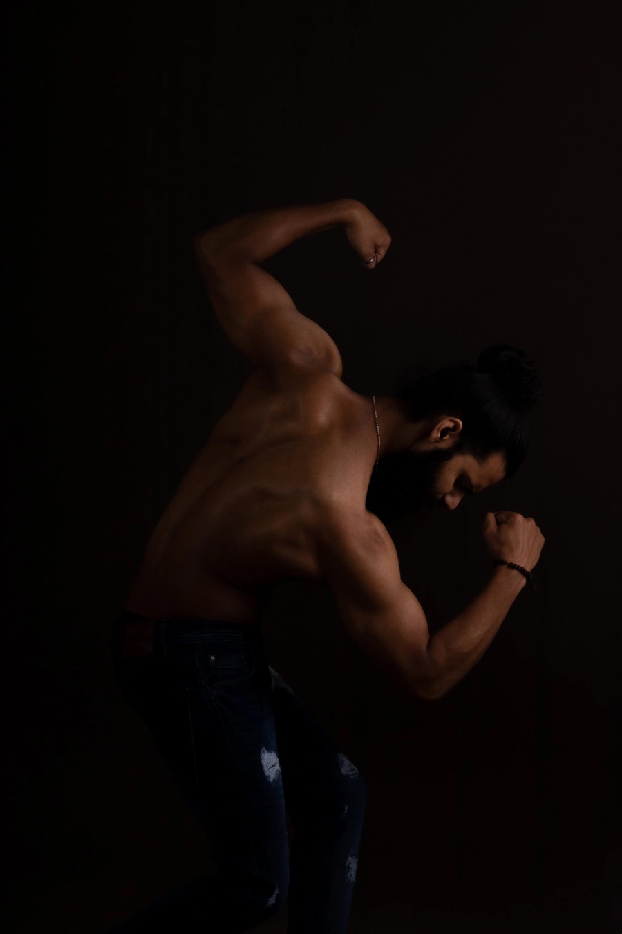 Shirtless physique study in low warm light, arm flexed and head turned away, deep brown backdrop
