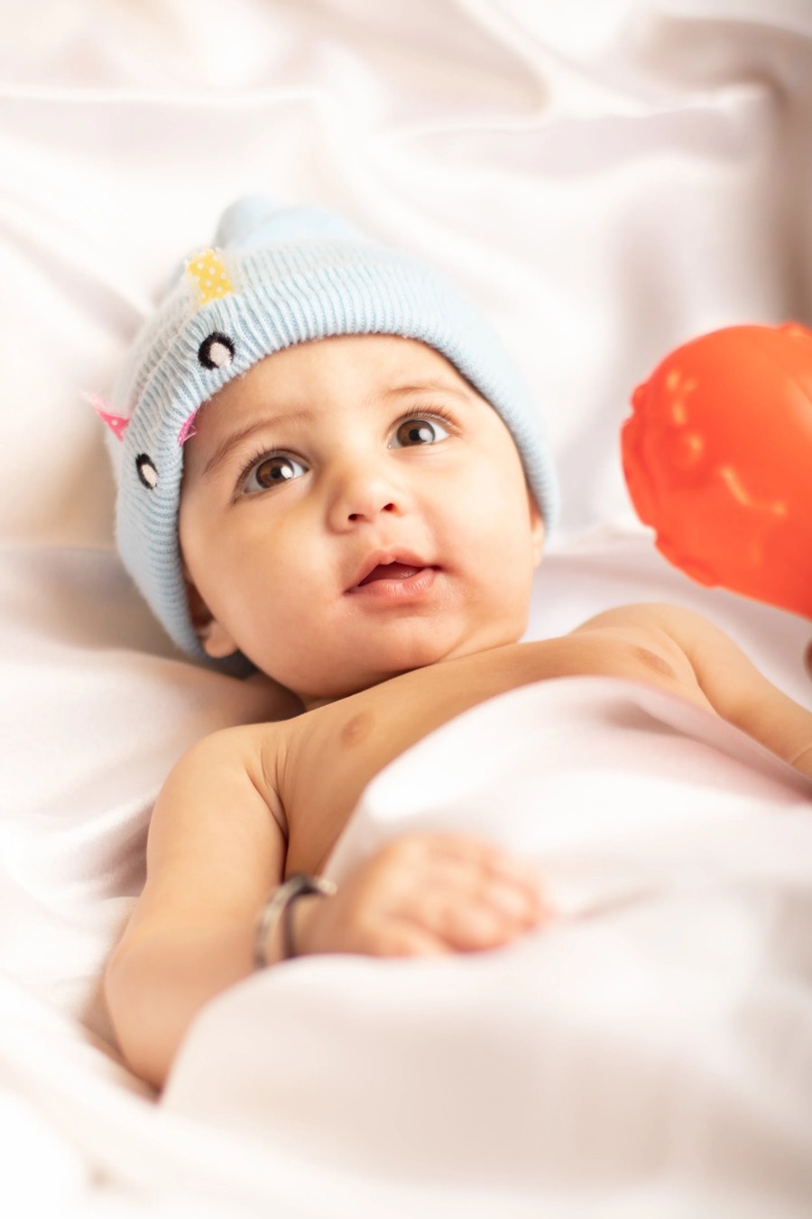 Baby in a pale blue knitted cap lying on white satin, orange ball at the edge of soft high-key light