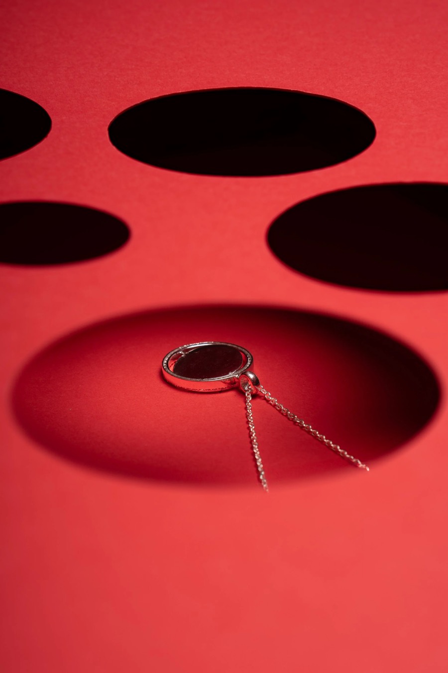 Round dark-stone pendant on a fine chain resting in an oval shadow cut into a red paper surface