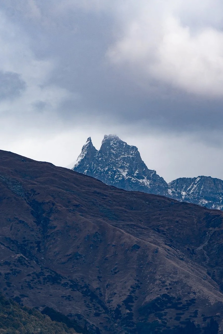 Twin rock summit dusted with snow breaking through cloud above a bare brown ridge, Waichin, Himachal