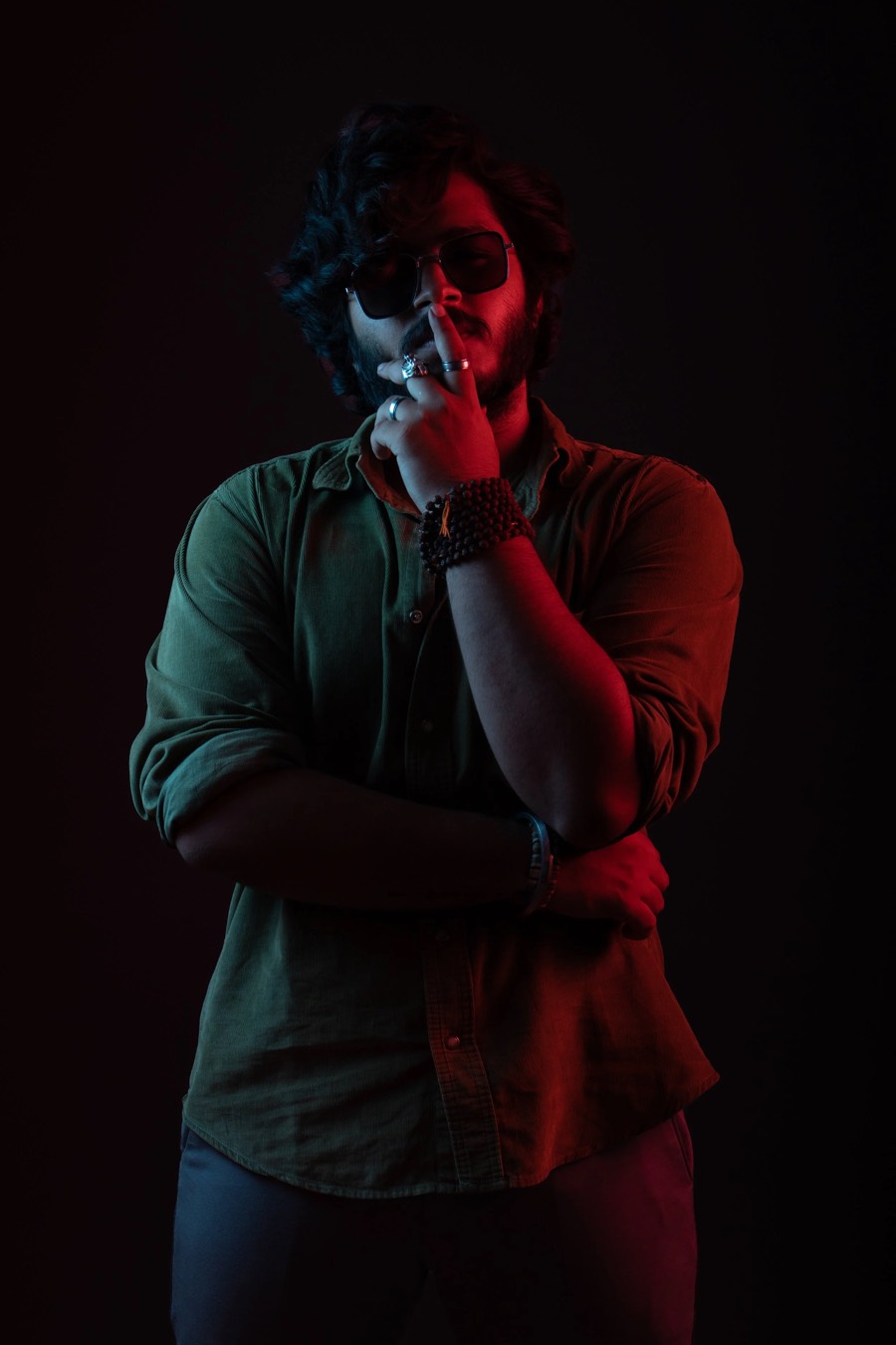 Red and blue gel lighting on a half-length frame, olive shirt and sunglasses, hand raised to the chin