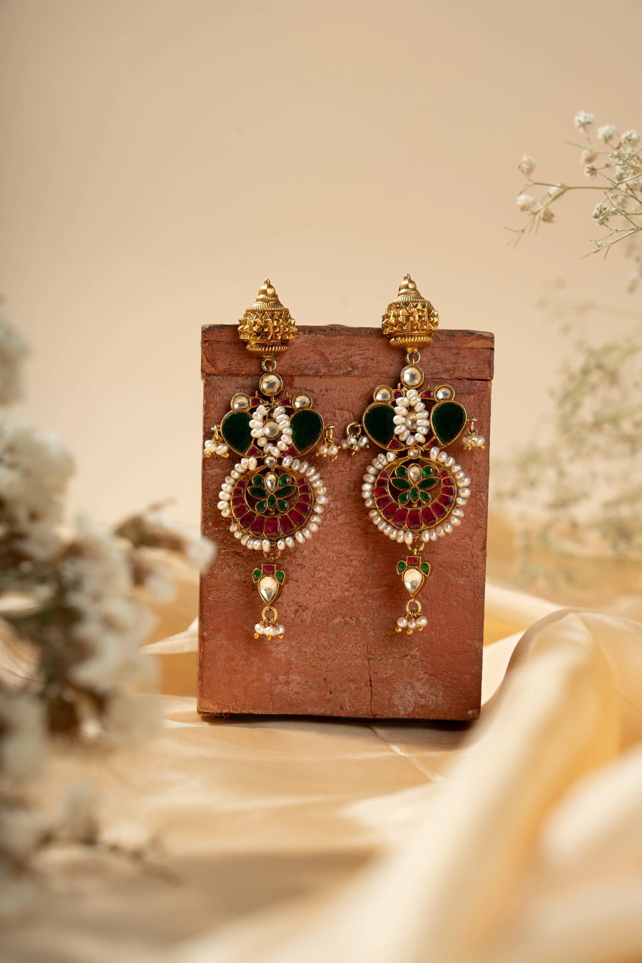 Chandelier earrings in red and green stones with pearl drops, propped against a terracotta block