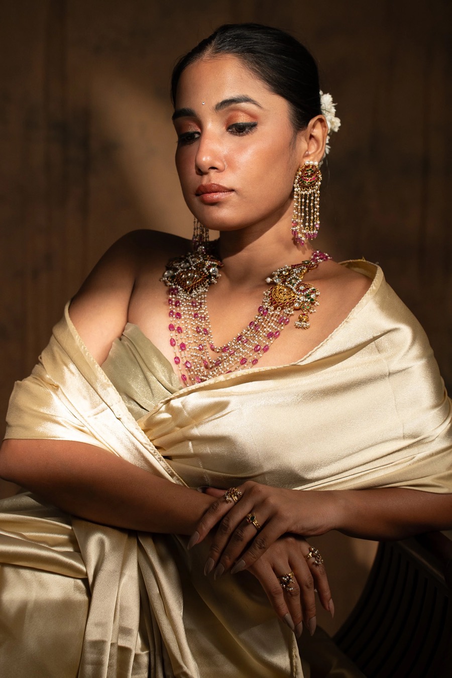 Seated in ivory satin with a layered stone necklace and long chandelier jhumkas, warm wood behind