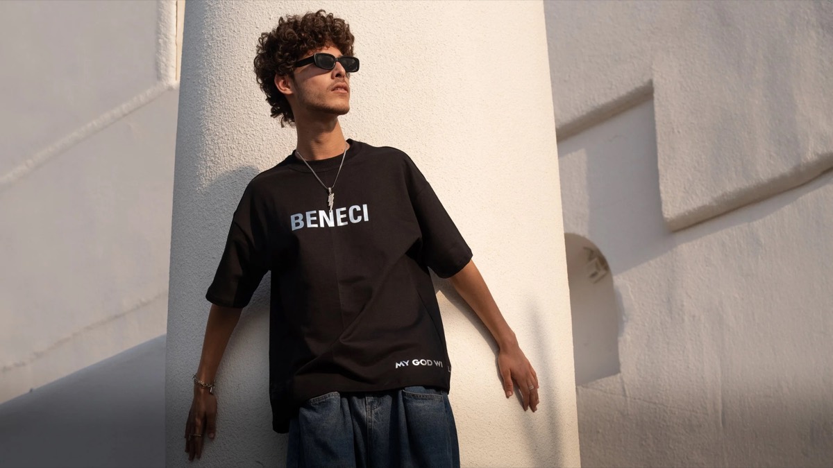 Model against a white wall in a black Beneci tee and sunglasses, hard midday sun raking across