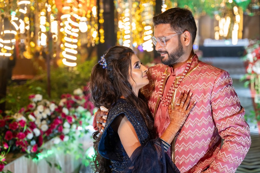 Evening portrait beside banked flowers, navy sequinned outfit and pink zigzag sherwani under string lights