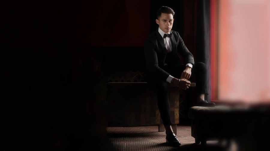 Full-length tuxedo frame in a wide dark room, foot up on a stool, pink-lit wall and red drape at right