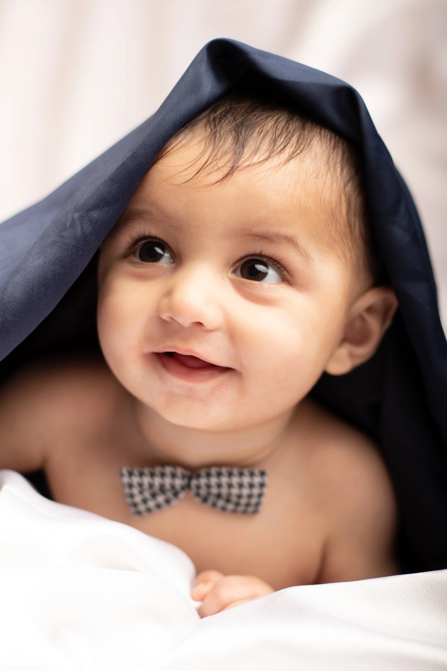 Smiling child peeking from under a navy drape, gingham bow tie and white bedding in bright soft light