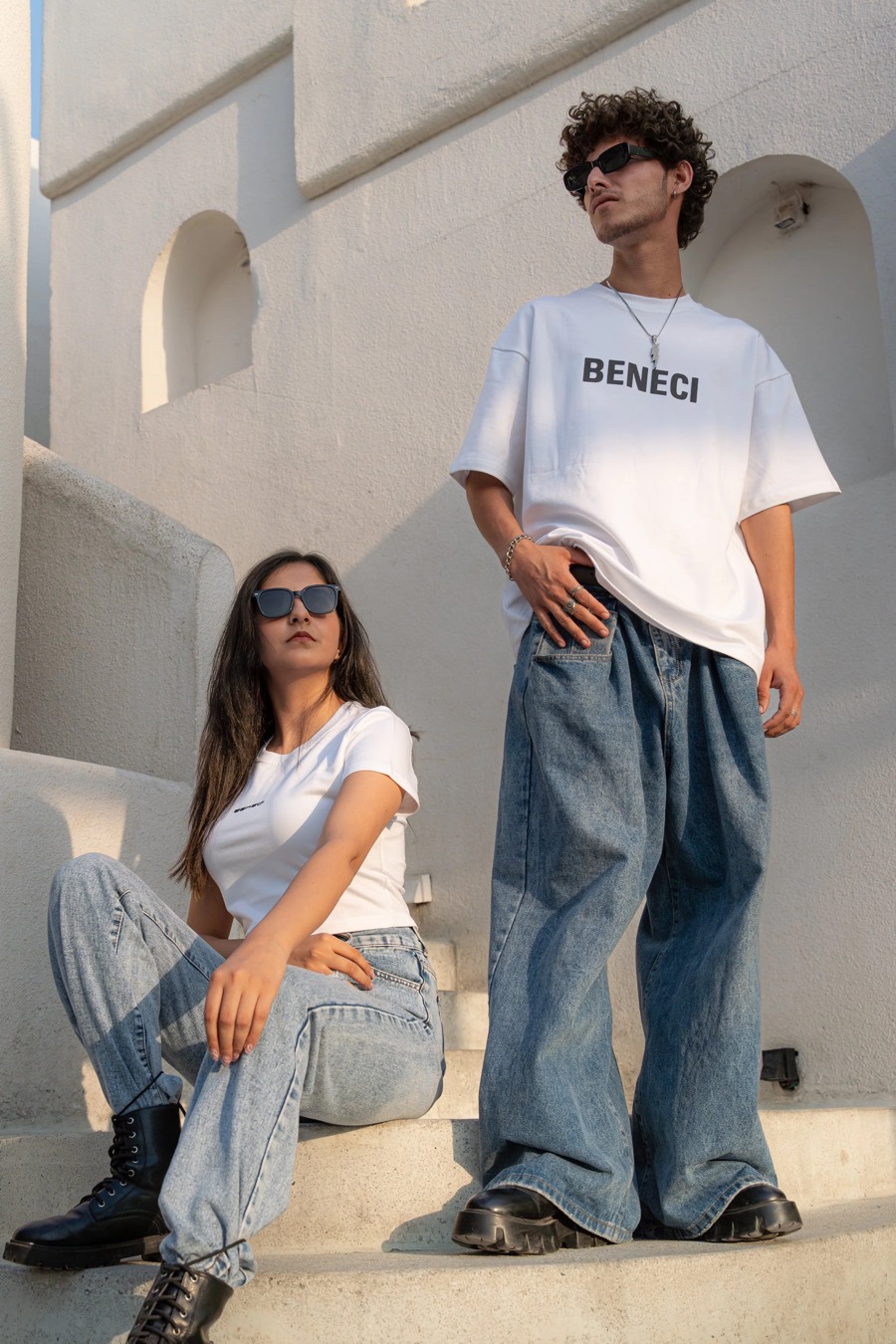 Two Beneci models on sunlit white steps, one standing in a logo tee, one seated in washed denim