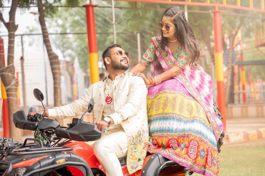 Groom in a cream sherwani astride a red motorcycle, bride in a pink ikat lehenga leaning in behind him
