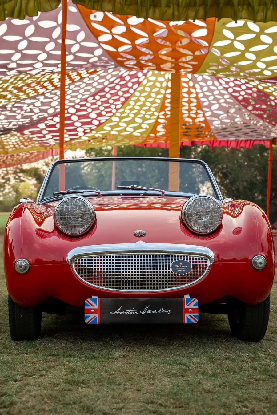 Small red roadster head-on under a patterned canopy, bonnet-top headlamps and a chrome oval grille