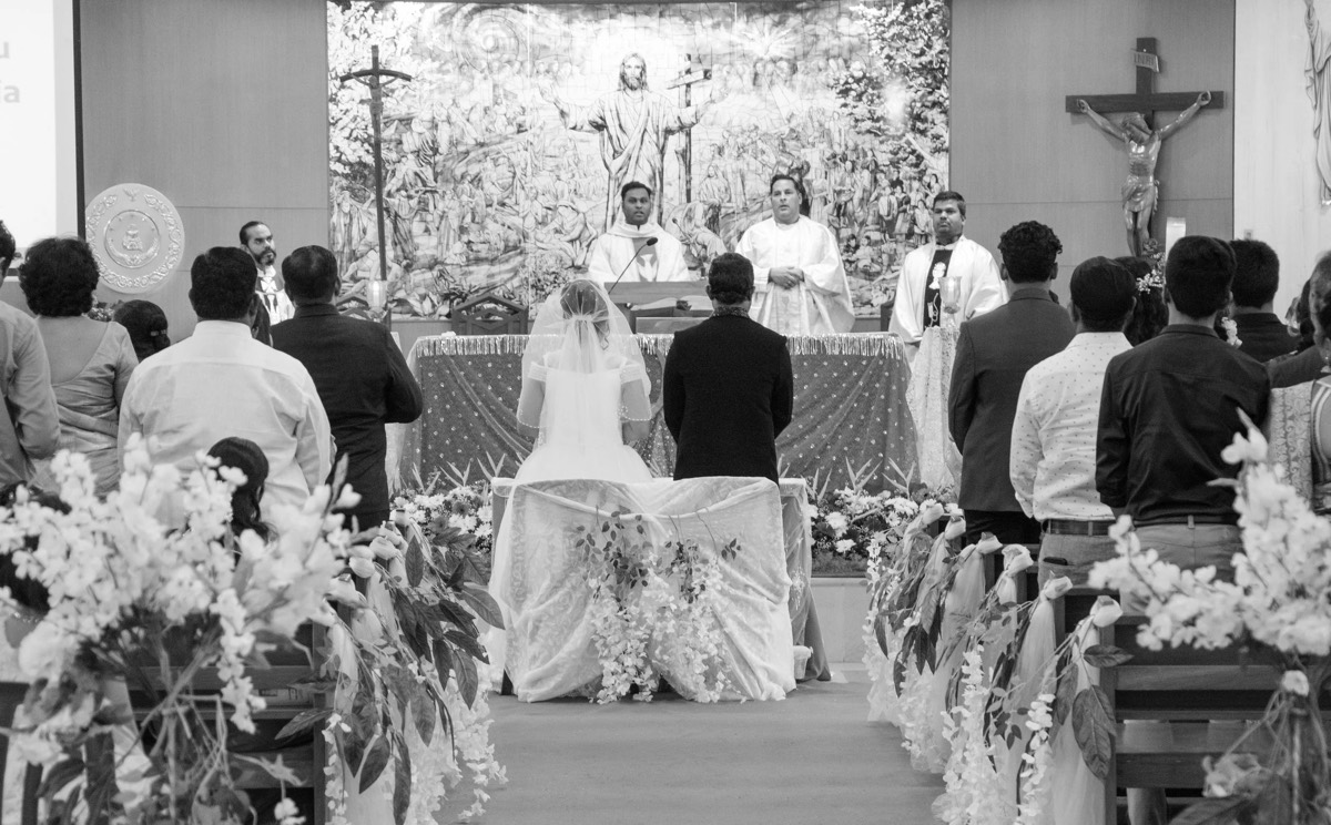 Black-and-white church wedding ceremony, the couple seated at the altar beside a flower-lined aisle