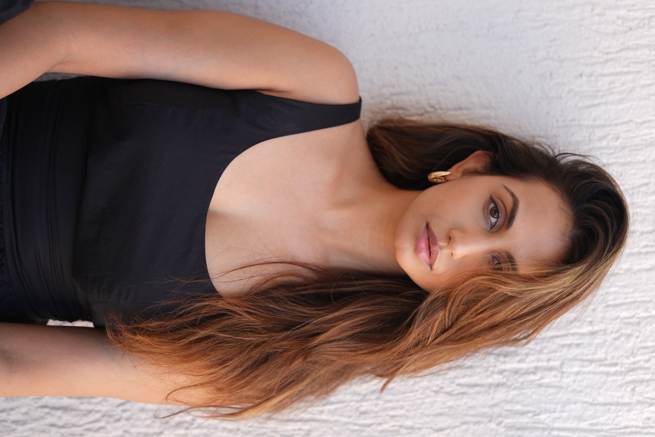 Anjali Schmuck against a white textured wall in a black scoop-neck top, direct gaze, hard daylight
