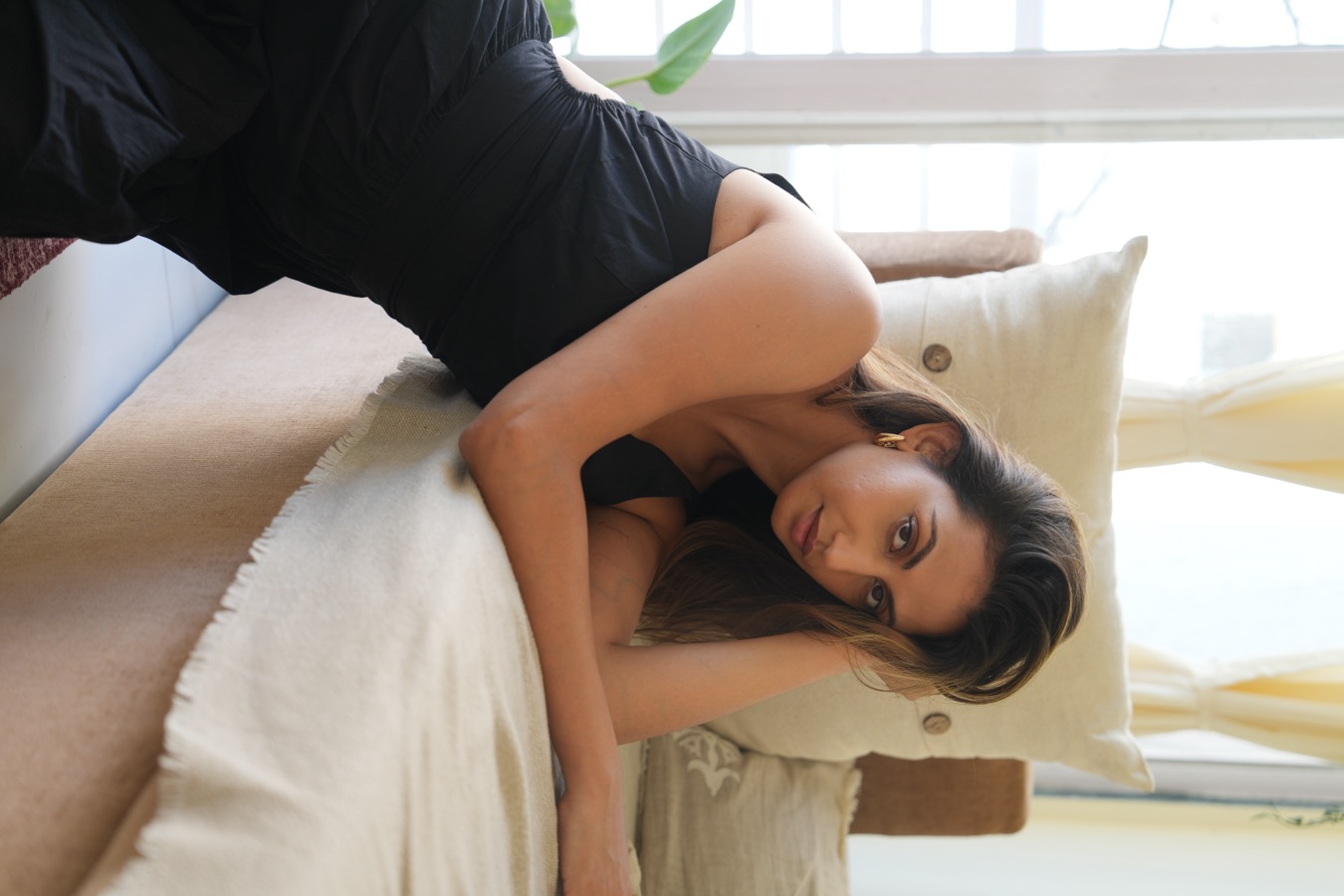 Leaning on a beige daybed cushion in a black dress, Anjali Schmuck holds the lens, window light behind