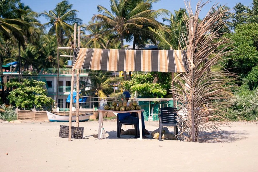 Striped awning shack with plastic chairs on white sand, palms and a fishing boat behind, Goa