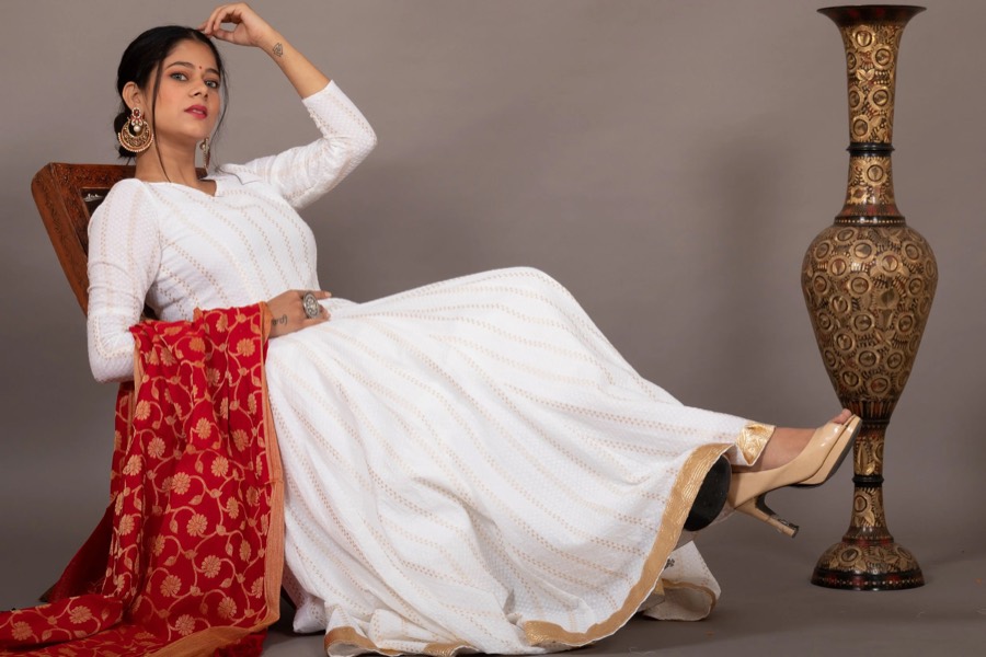 White floor-length anarkali with red brocade dupatta, model reclining beside a tall brass vase