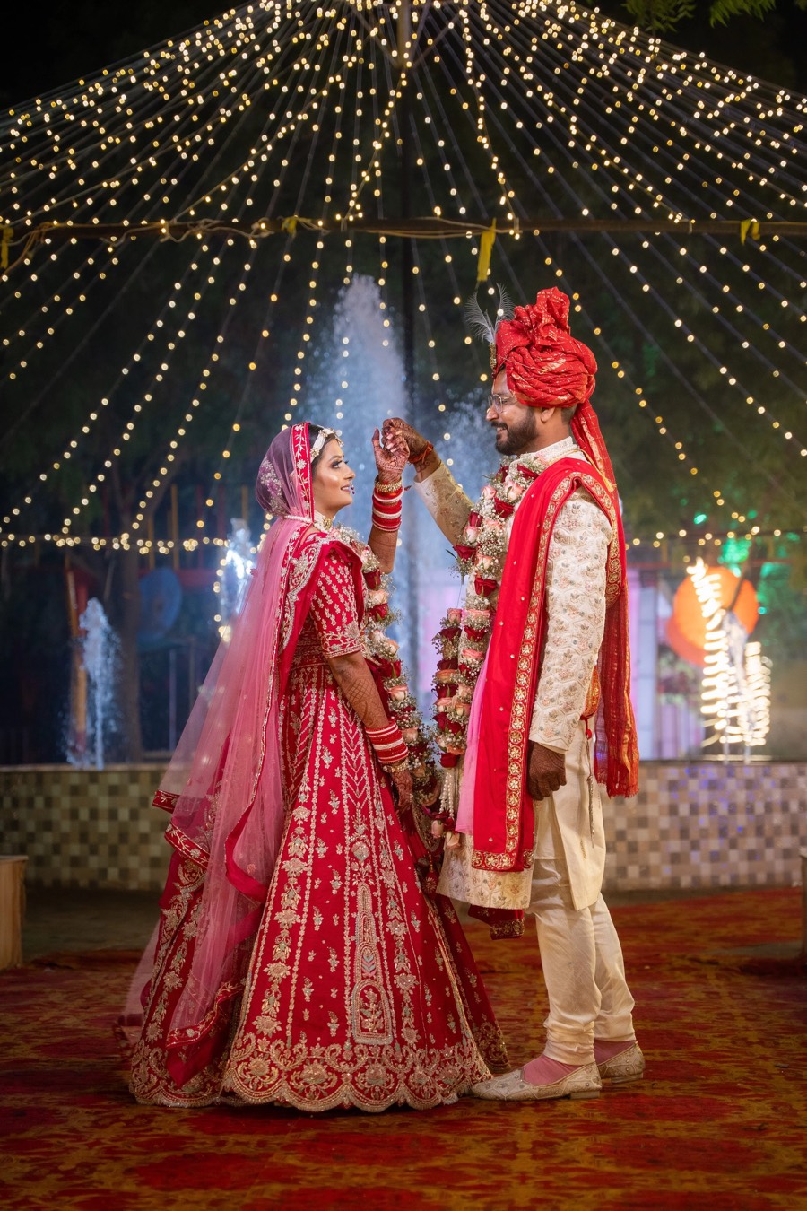 Red lehenga and red turban, hands joined and lifted under a canopy of fairy lights on a patterned carpet
