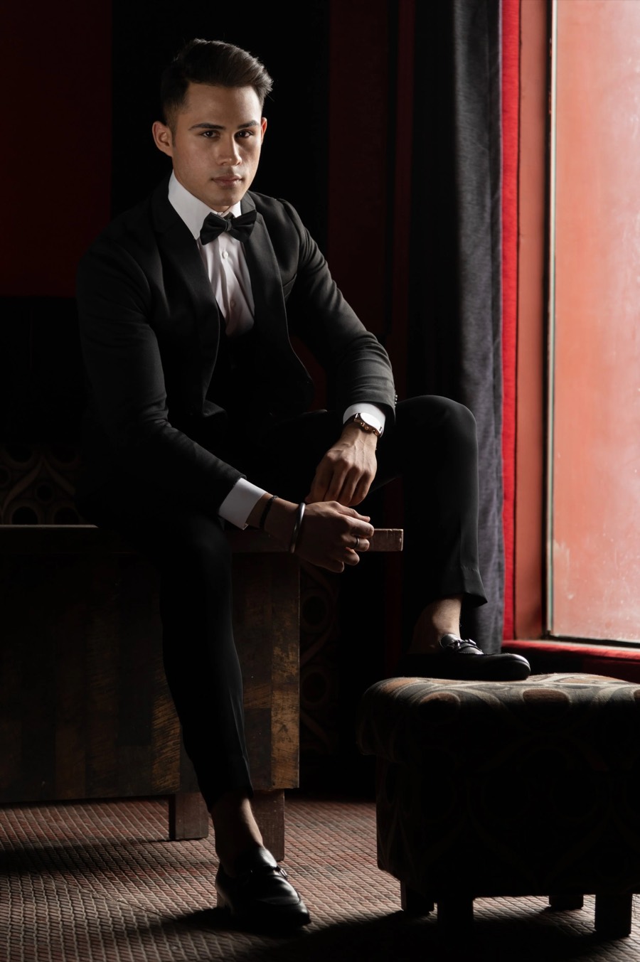 Vertical full-length in a black tuxedo, leaning forward with hands together beside a deep red curtain