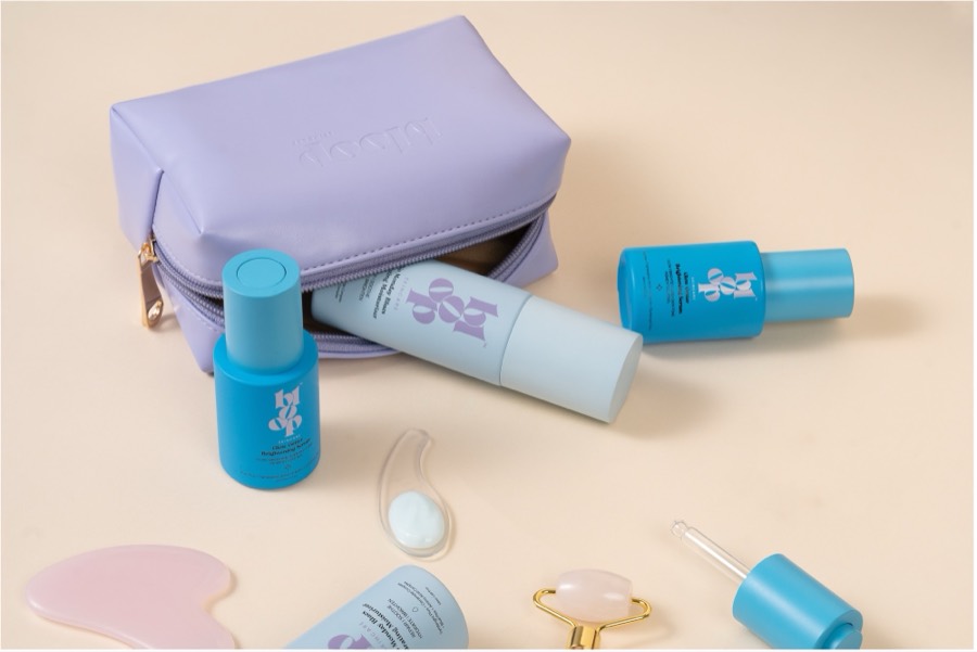 Lilac zip pouch with blue Bloop bottles, a white tube, gua sha stone and roller laid out on beige