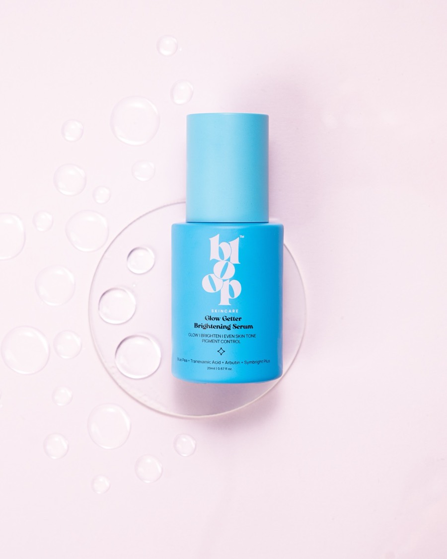 Glow Getter Brightening Serum in cobalt glass on a clear disc, water beads scattered across soft pink
