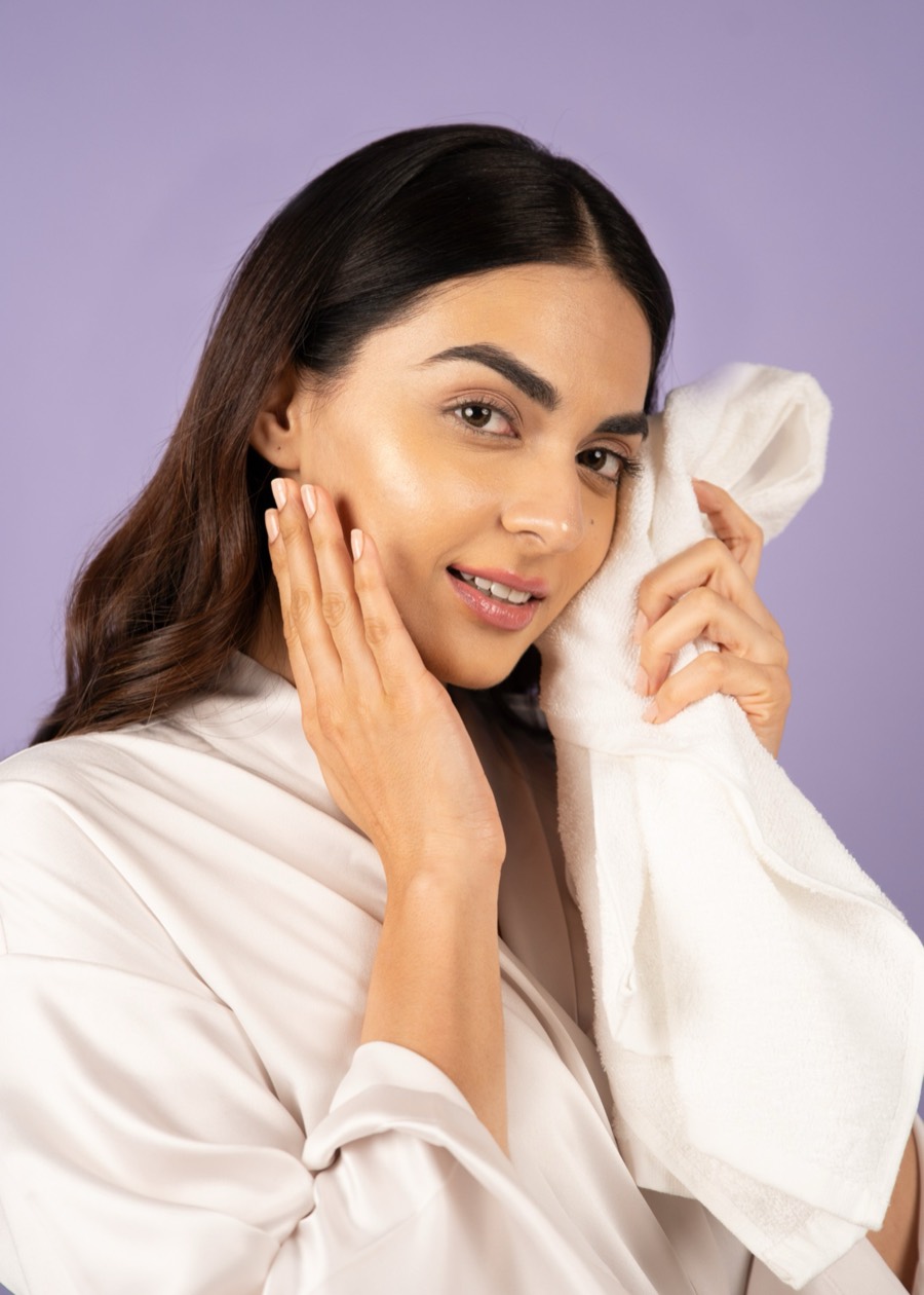 Model in a white shirt pressing a folded towel to her cheek against a lilac studio backdrop