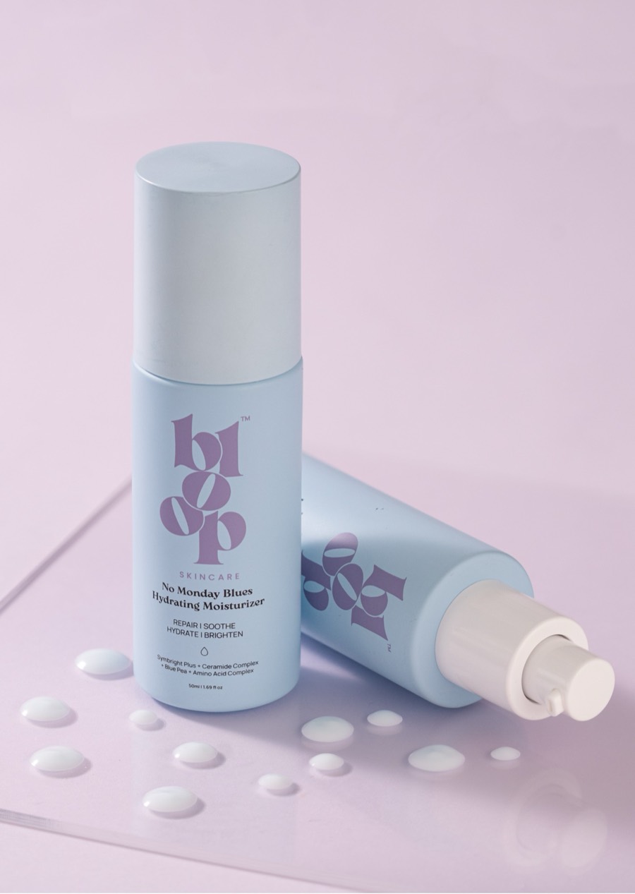 Two pale blue Bloop moisturisers, one upright and one on its side, cream droplets on a pink plinth