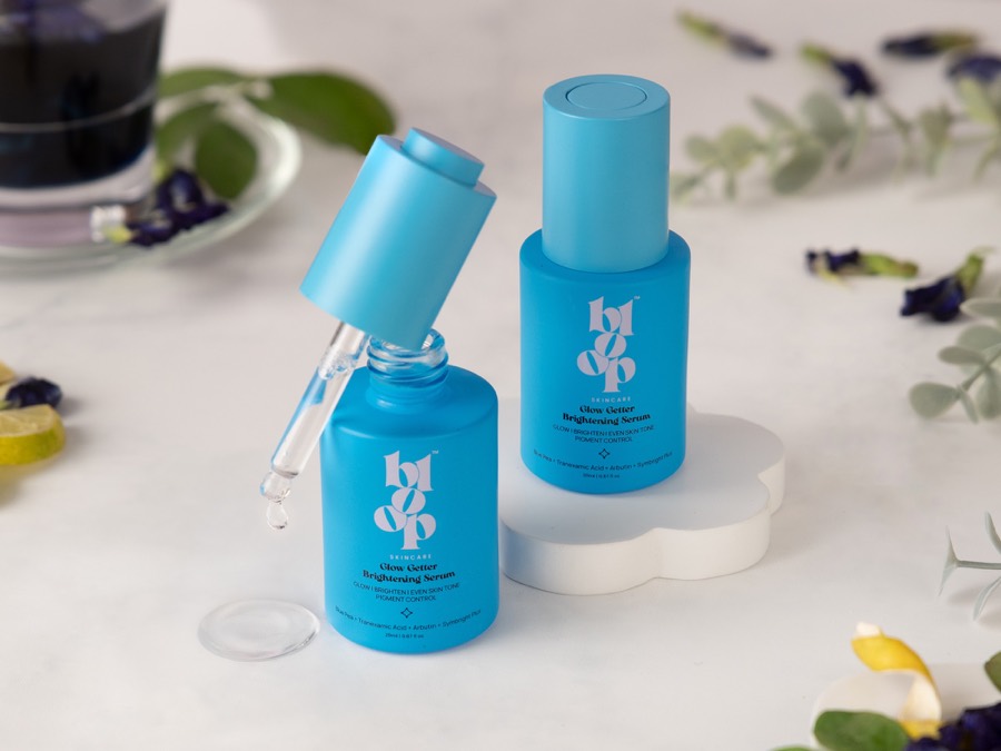 Cobalt serum pair on white marble, dropper lifted with a drip forming beside petals and dark tea