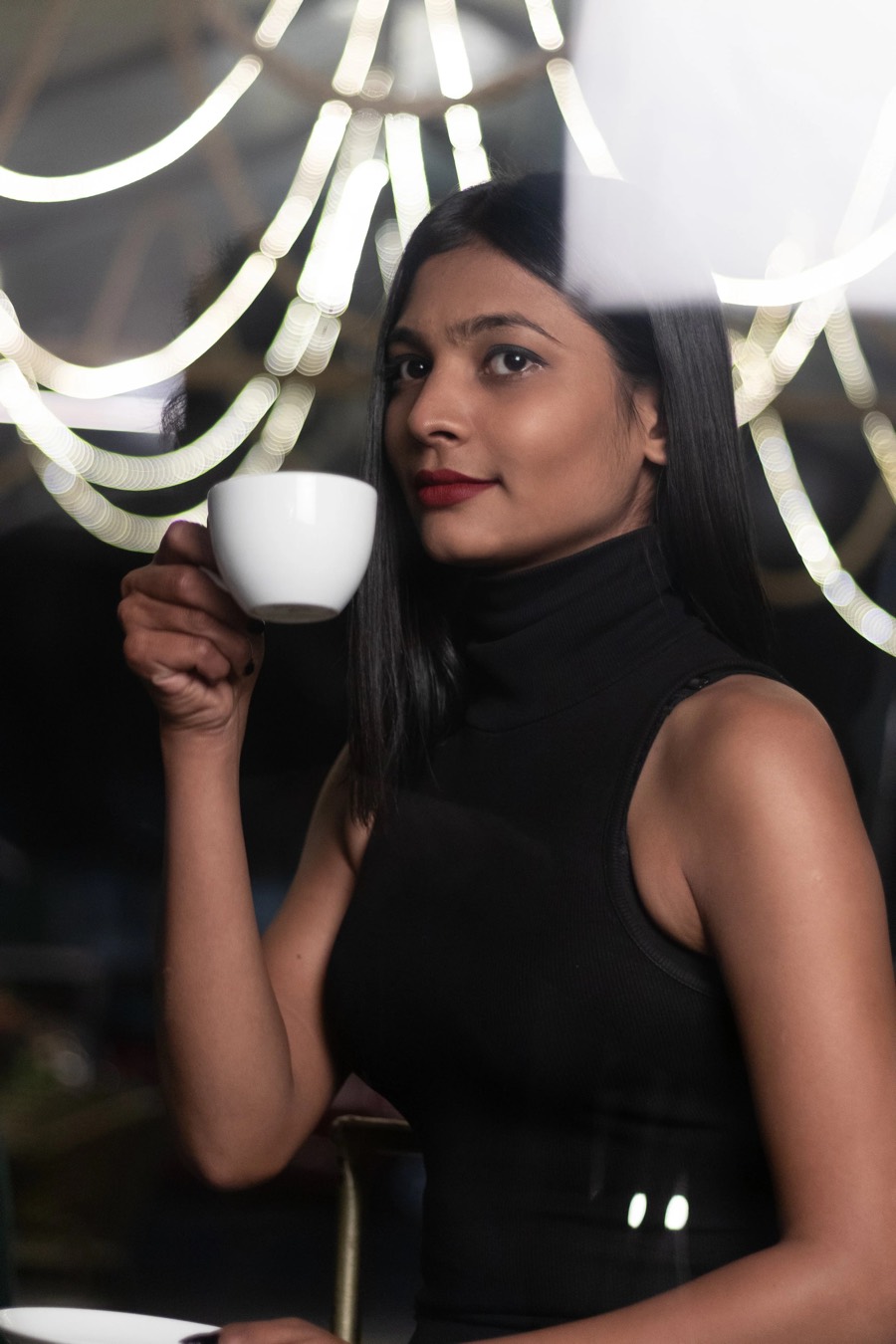 Black sleeveless turtleneck and a white cup raised, café bokeh behind, portrait by Curtain Drop Media
