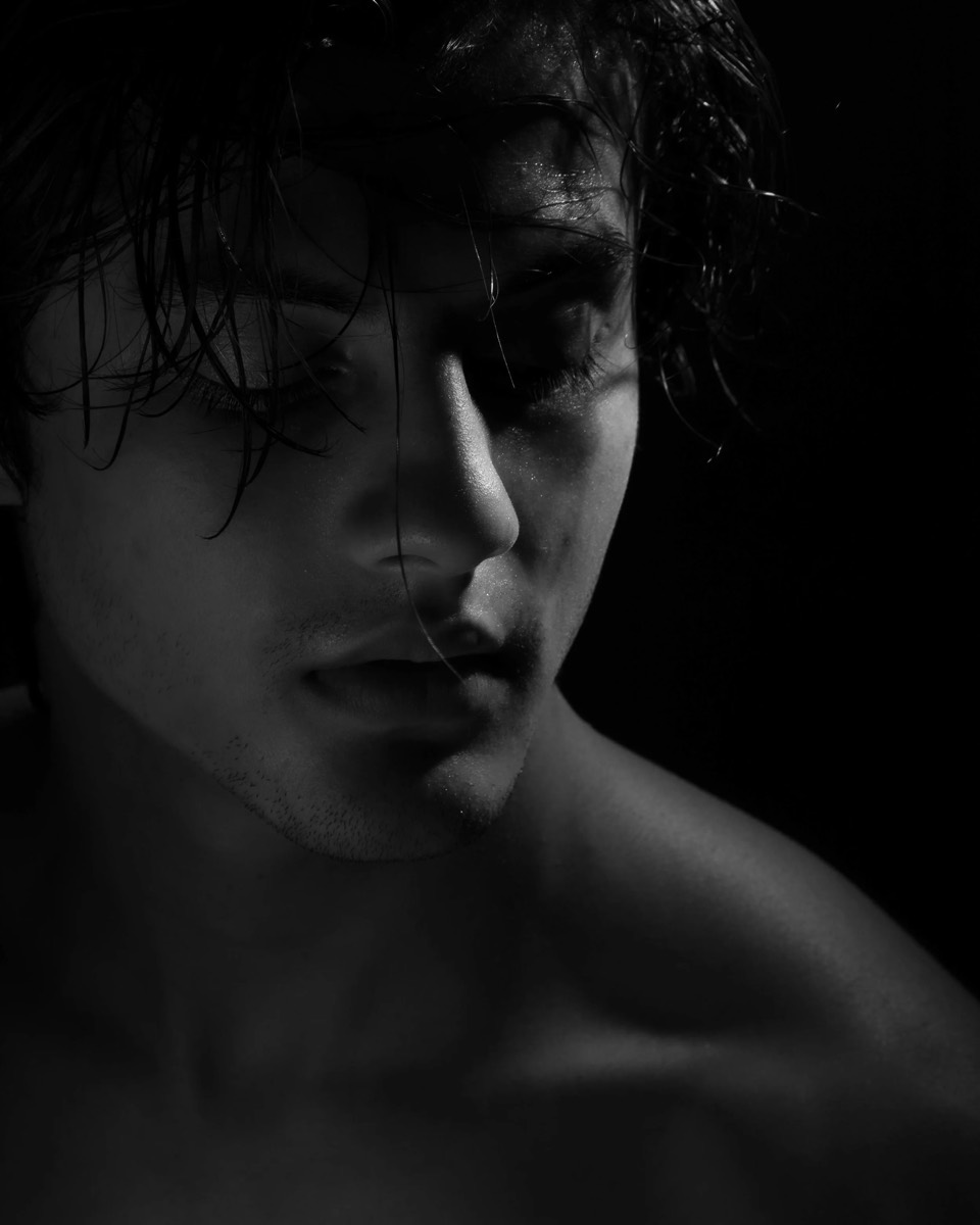 Low-key black and white close-up, damp tousled hair and bare shoulder emerging from near-total shadow