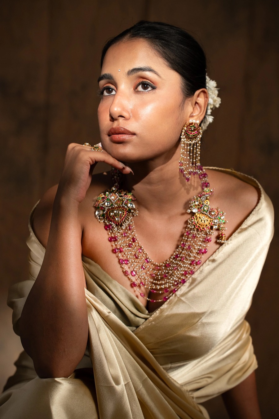 Hand to chin in ivory satin, a long pink-beaded haar and kundan pendant against a brown backdrop