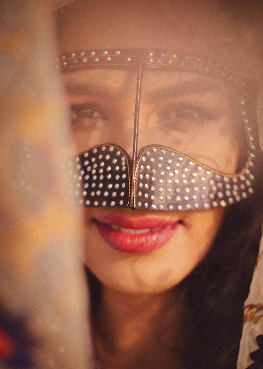 Warm hazy close-up shot through fabric, an ornate studded veil across the face and a red-lipped smile