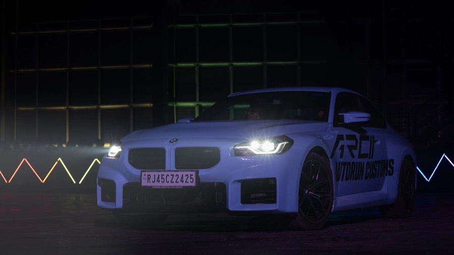 White coupe with motorsport decals on the door, headlights blazing in a dark hall, light trails behind