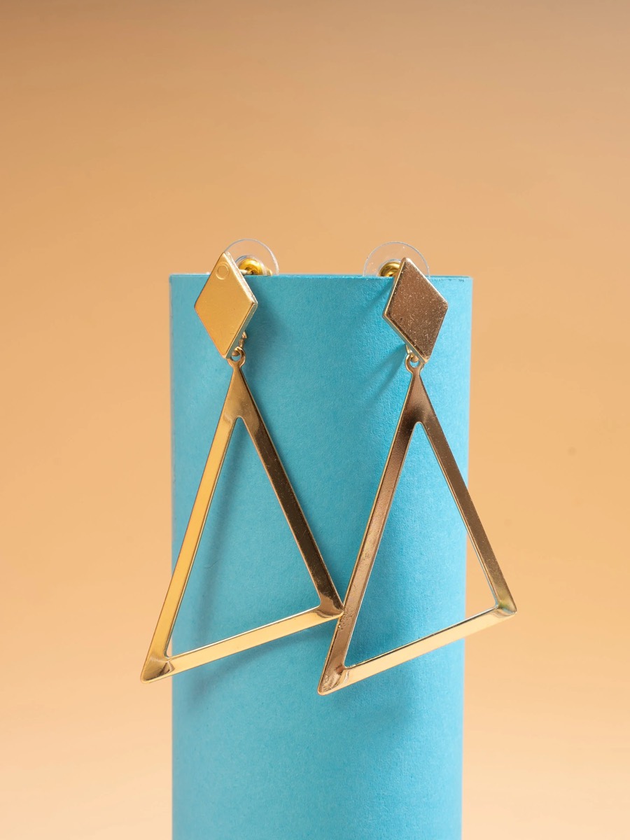 Geometric gold triangle drop earrings hung on a teal block against a tan backdrop for Viraasi