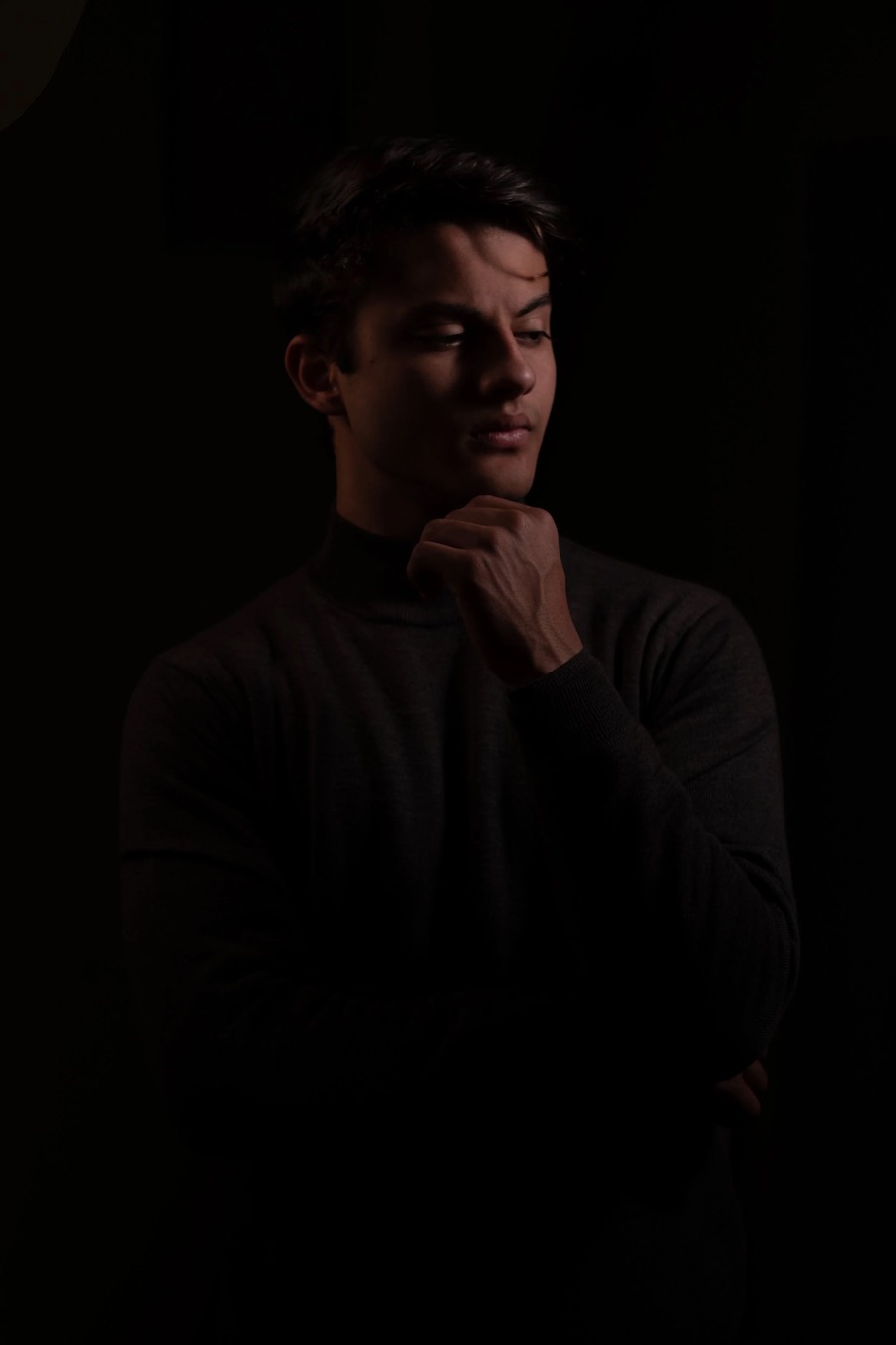 Dark turtleneck and folded arms, a single dim light catching the jaw, head turned into deep shadow
