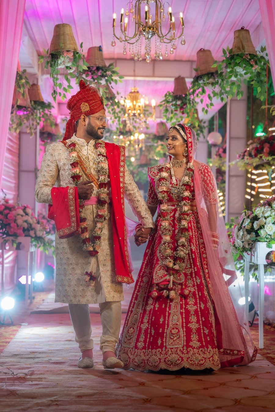 Hand in hand through a floral-draped entrance lit by a chandelier, red bridal lehenga and cream sherwani