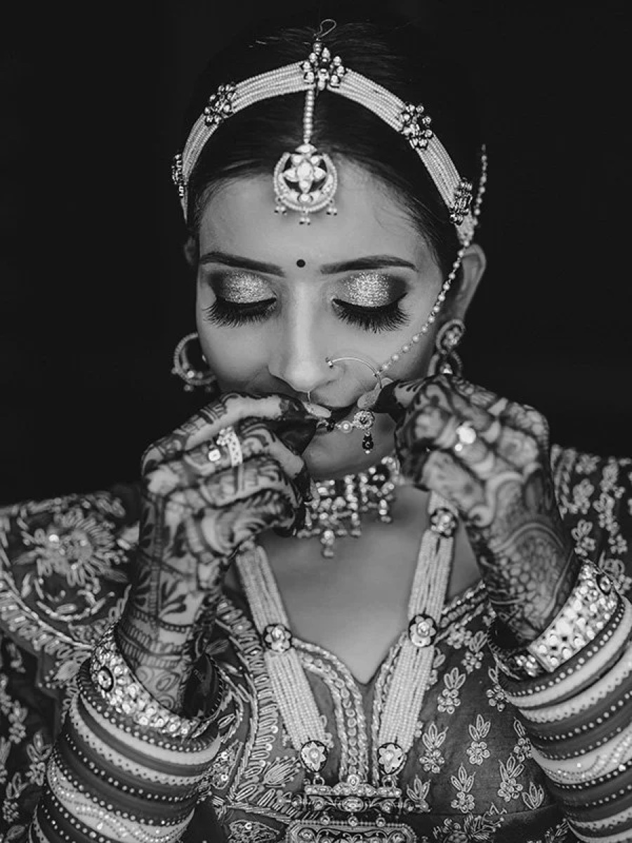 Monochrome bridal portrait with mehendi-covered hands raised to the face, eyes lowered, nose-ring chain
