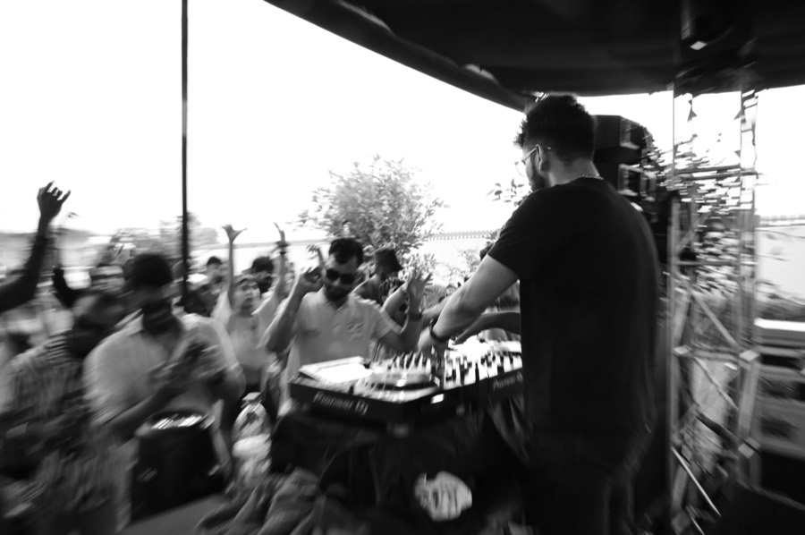 Black and white frame of a DJ at the decks facing a crowd with hands up, open-air stage in Jaipur