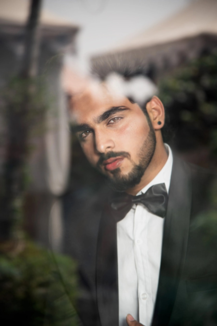 Shot through glass with reflections across it, tuxedo and bow tie, hand at the lapel, gaze to camera
