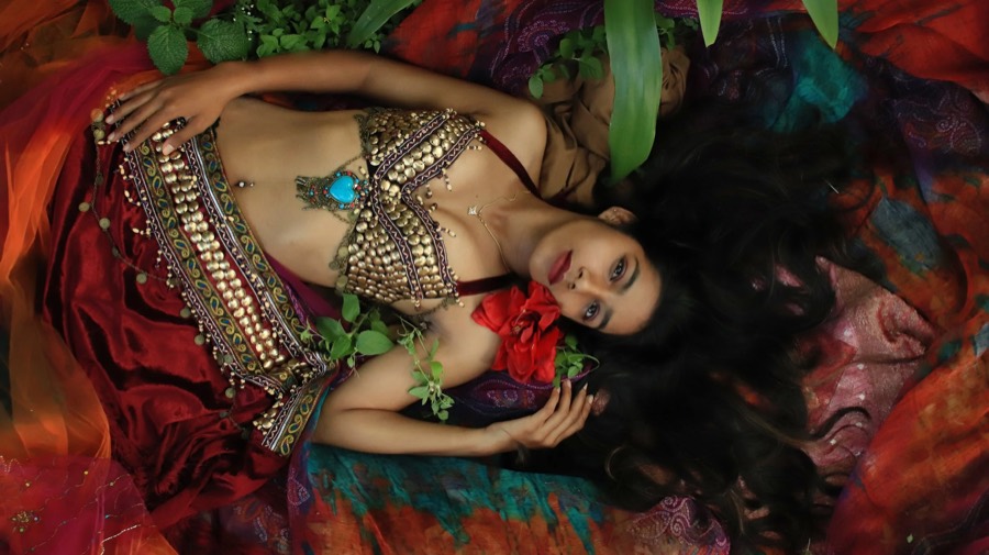 Lying on layered red and orange drapes in a gold coin bodice, hair fanned out, red bloom in hand