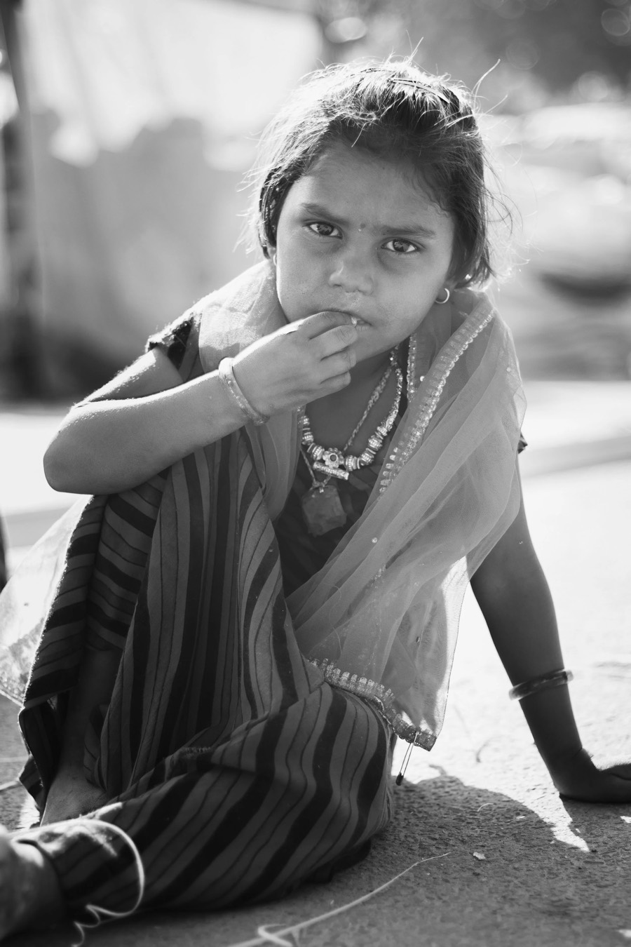 Seated girl in a striped wrap and light dupatta, beaded necklace and bangles, backlit black and white