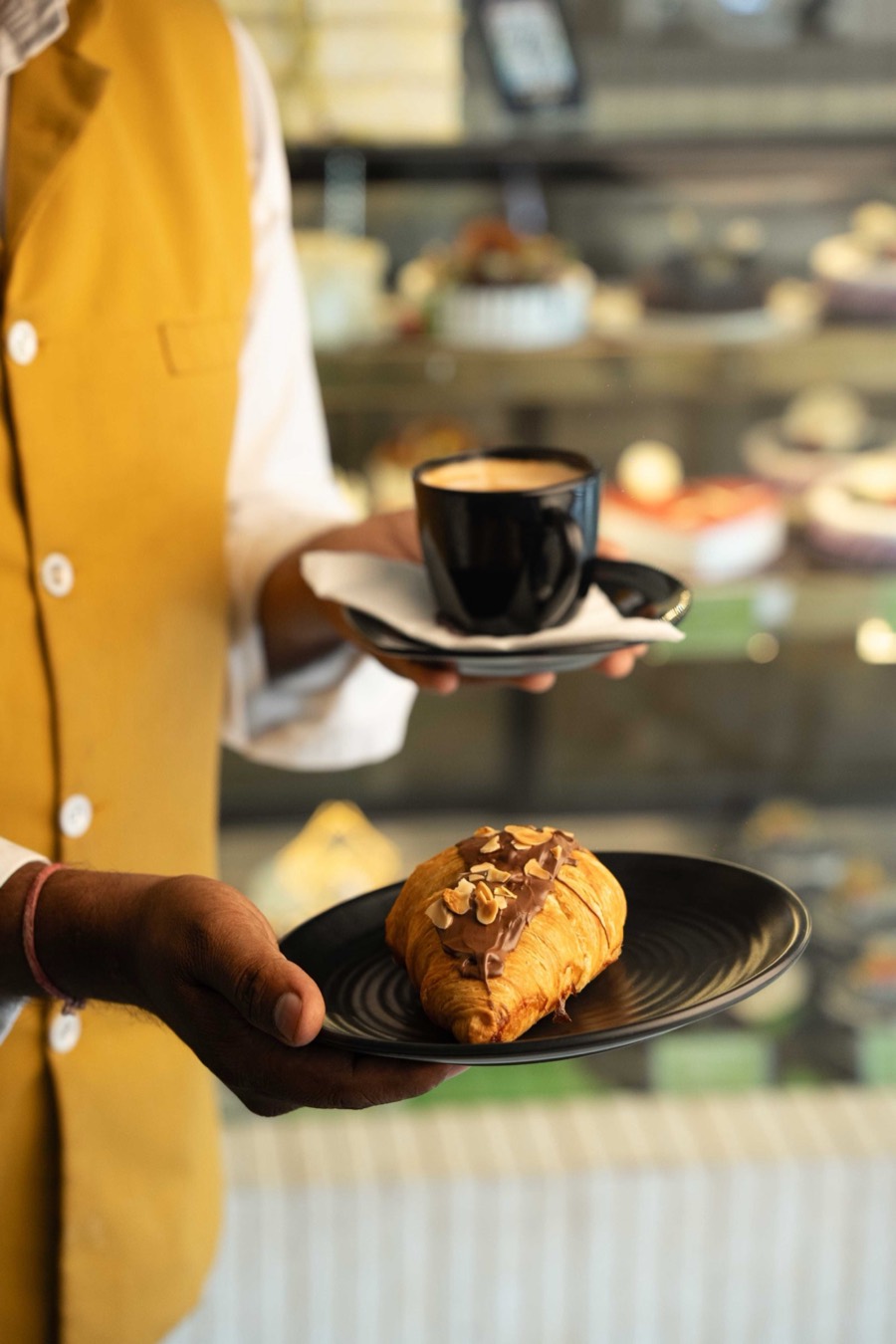 Server in a mustard waistcoat carrying a black coffee cup and a nut-topped croissant on two plates