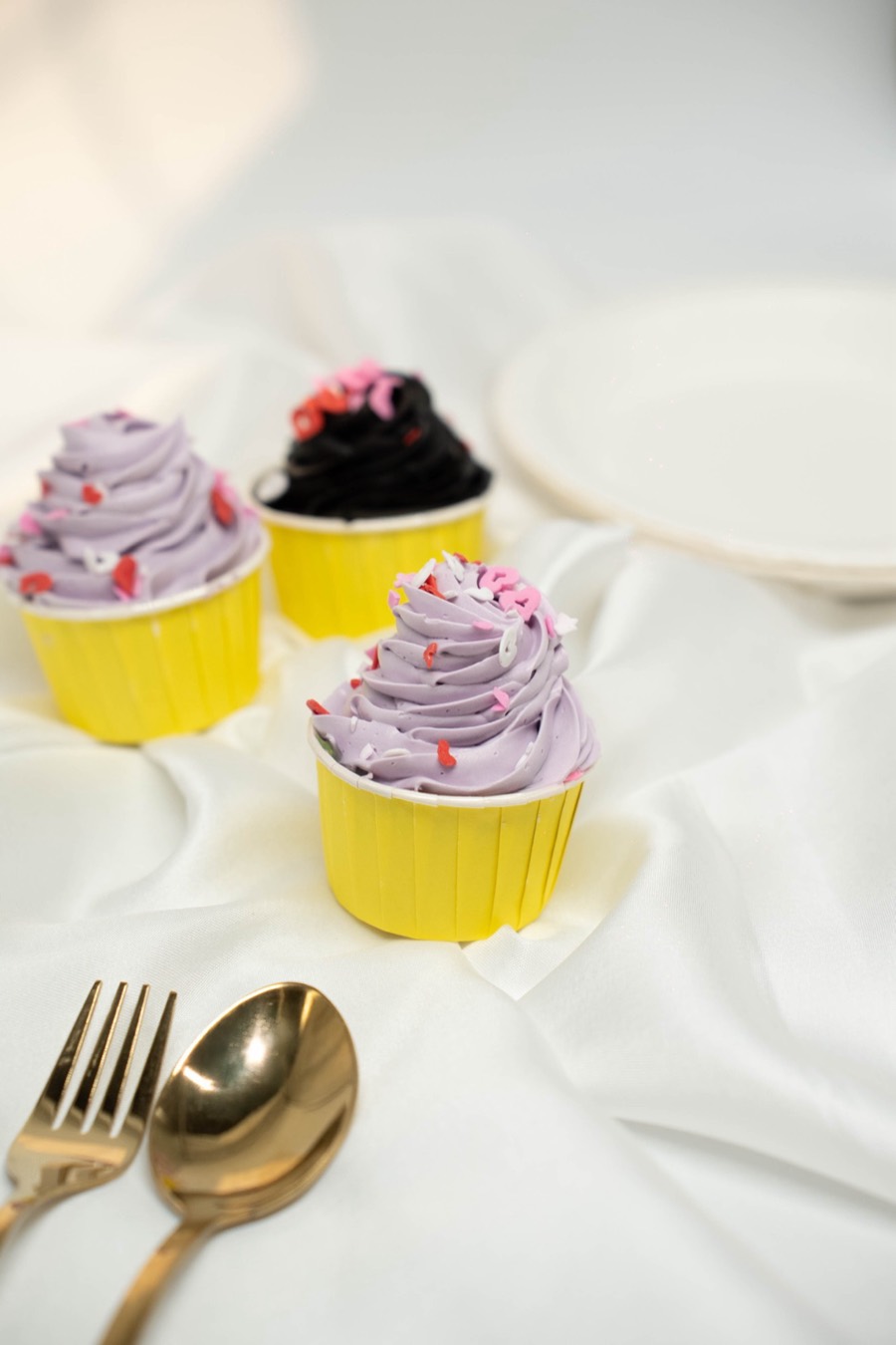 Cupcakes in yellow cases with lilac swirled icing and pink sprinkles, gold fork and spoon alongside