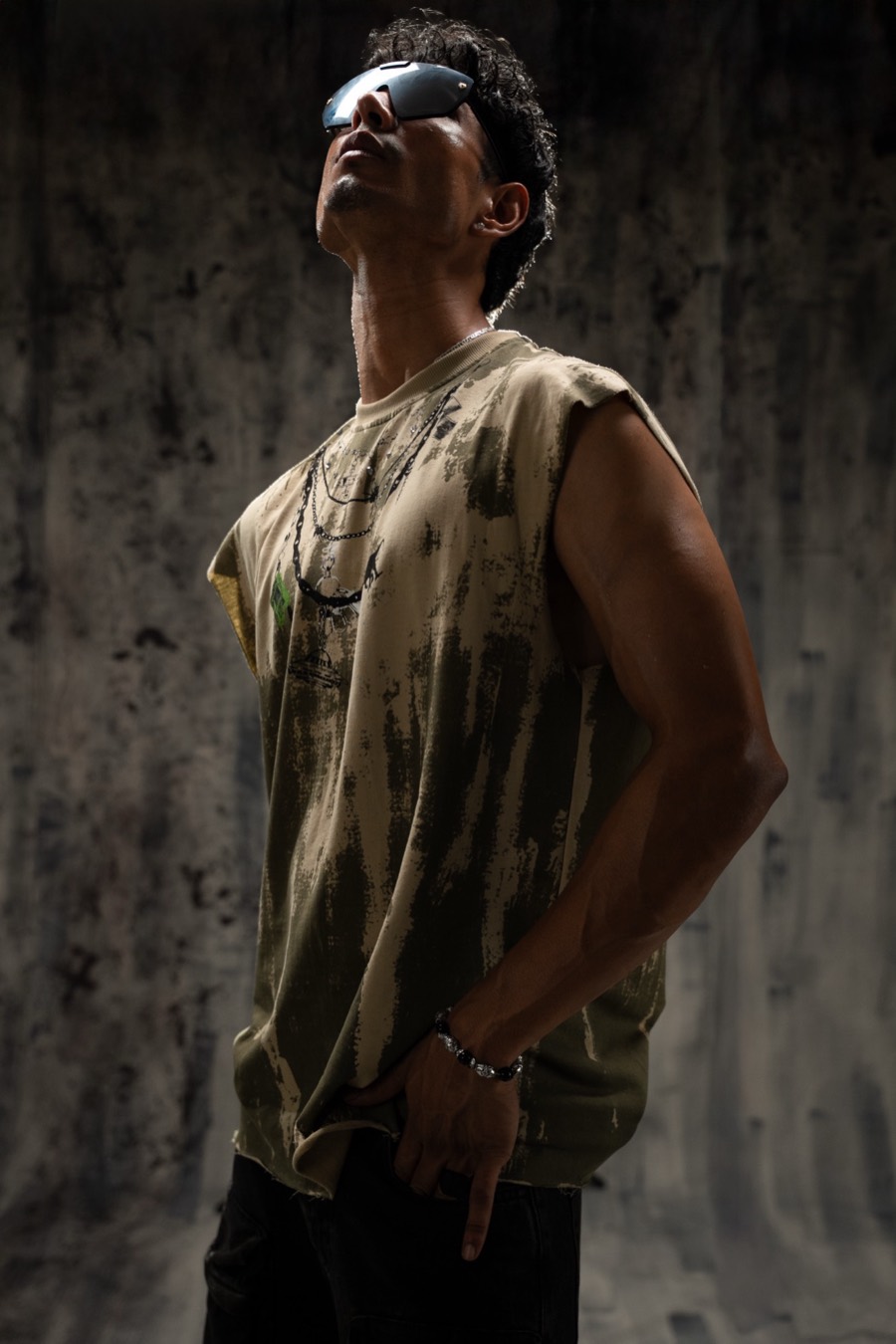 Distressed khaki sleeveless tee and wraparound shades, chin lifted against a dark concrete wall