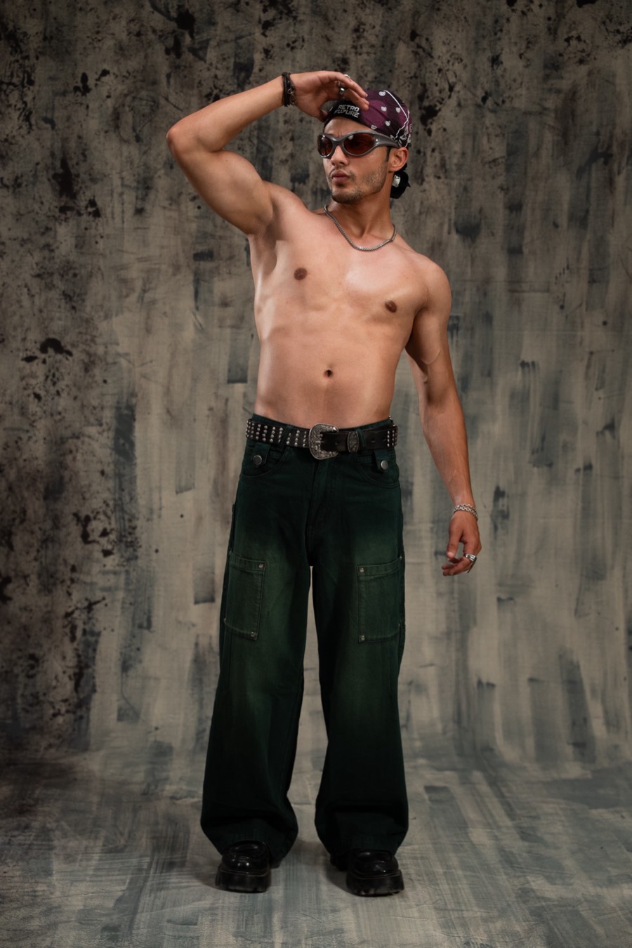 Shirtless full-length pose in dark green baggy cargo jeans, bandana tied back, one arm raised high