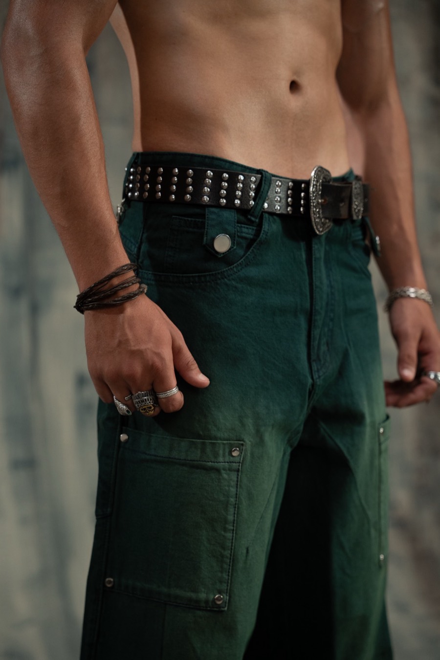 Waist crop of green cargo jeans with a studded belt, rings and stacked leather bracelets on both hands