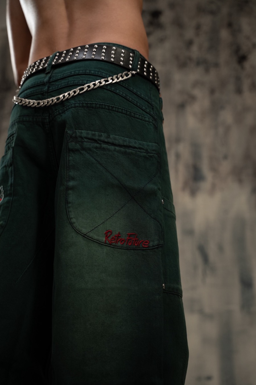 Back pocket detail of green jeans with red script embroidery, studded belt and hanging silver chain