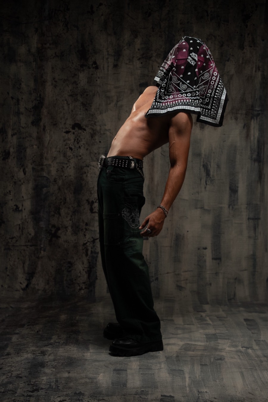 Bent-forward pose with a patterned bandana falling over the face, baggy black jeans and chunky boots
